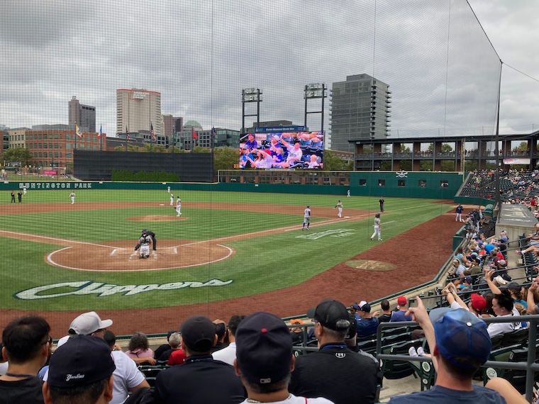 A baseball field as seen from the stands behind home plate. Players occupy their regular positions at the bases. Aside from one player walking back to the home team’s dugout at right, the area of the field in front of the dugout is empty.