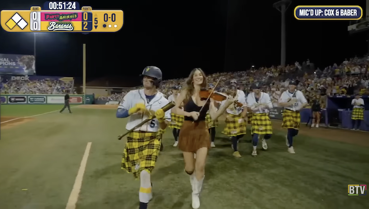 Screenshot from livestreamed Banana Ball game, showing a parade of players in the area in front of the home team’s dugout. The batter, wearing a yellow kilt, uniform jersey, and batting helmet, holds his bat in “air guitar” position; walking in step with him is a woman in a tank top, short skirt, and cowboy boots, playing the fiddle. Following behind them is a group of seven more players in kilts, clapping in time to the music.