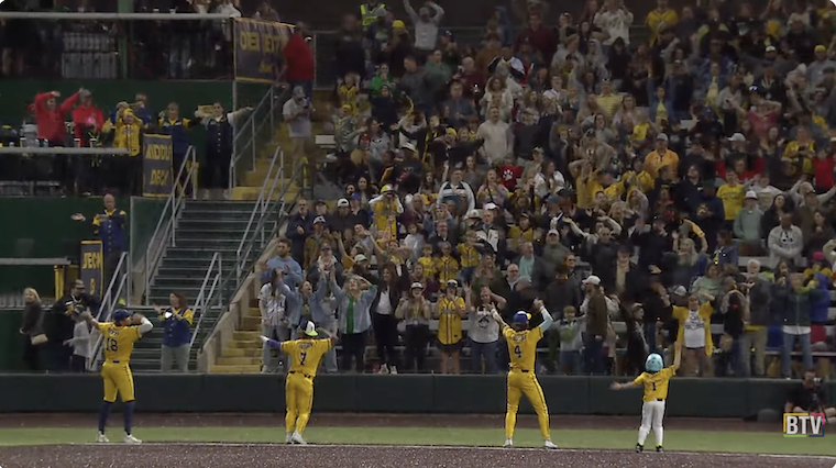 In the foreground, three uniformed Savannah Bananas players and one young fan in a Bananas jersey are seen from the back, standing on the field along the baseline, facing the crowd in the stands. Their hands are raised over their heads and they are dancing. Members of the crowd in the stands are standing with their arms raised, mimicking the players’ gestures.