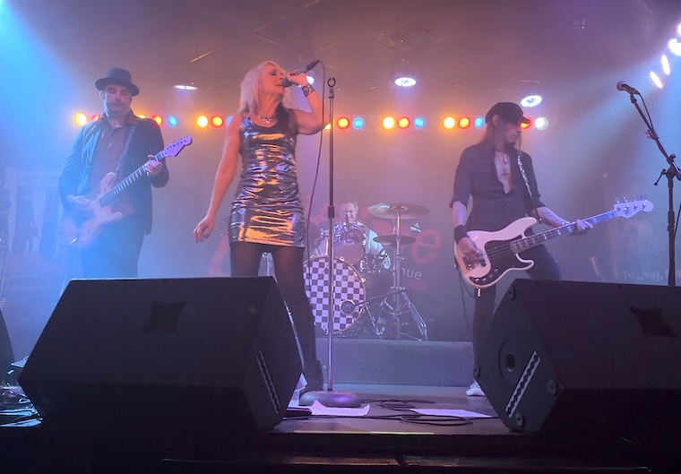 A photograph of a live band performing on a dimly lit stage. In the foreground, two large black stage monitors sit on the floor. On stage, a female vocalist in a shiny silver dress stands center stage at a microphone stand. To her left, a guitarist wearing a fedora plays a light-colored electric guitar. To her right, a bassist in black plays a white bass guitar. In the background, partially obscured, a drummer is seated at a drum kit with a black-and-white checkered bass drum head. The stage is illuminated by spotlights in shades of blue, orange, and red, creating a hazy, energetic atmosphere.